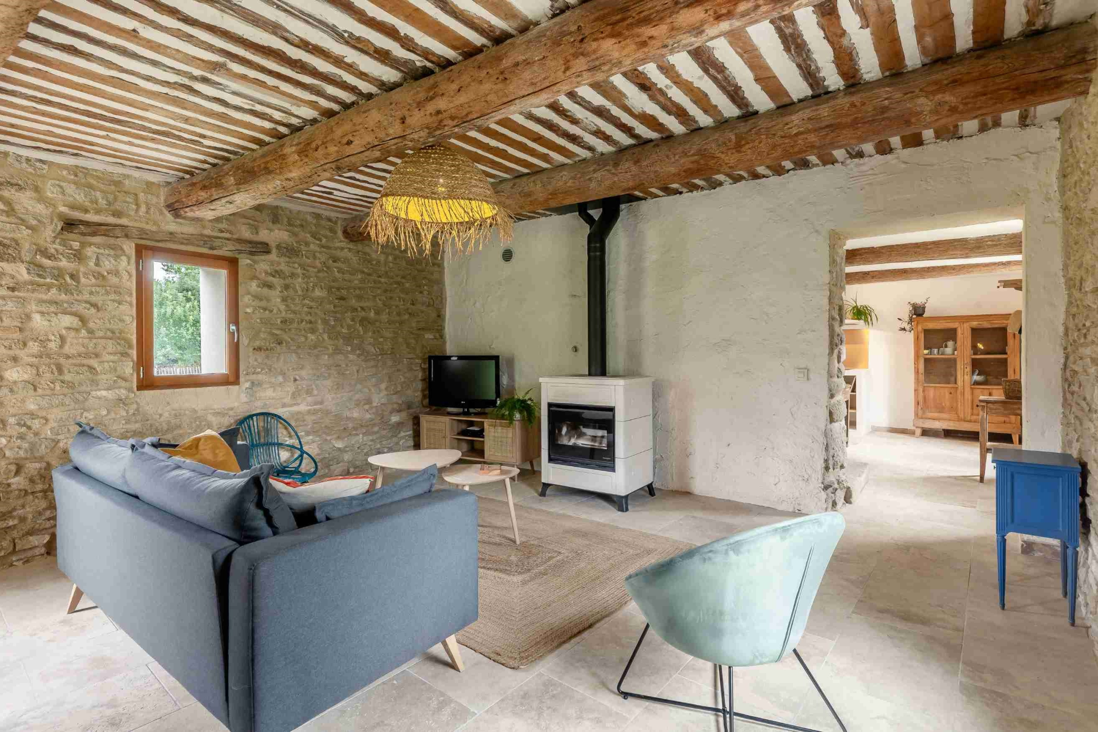Un salon rustique avec un plafond à poutres apparentes, des murs en pierre, un canapé bleu, un poêle à bois moderne, et un sol carrelé.
