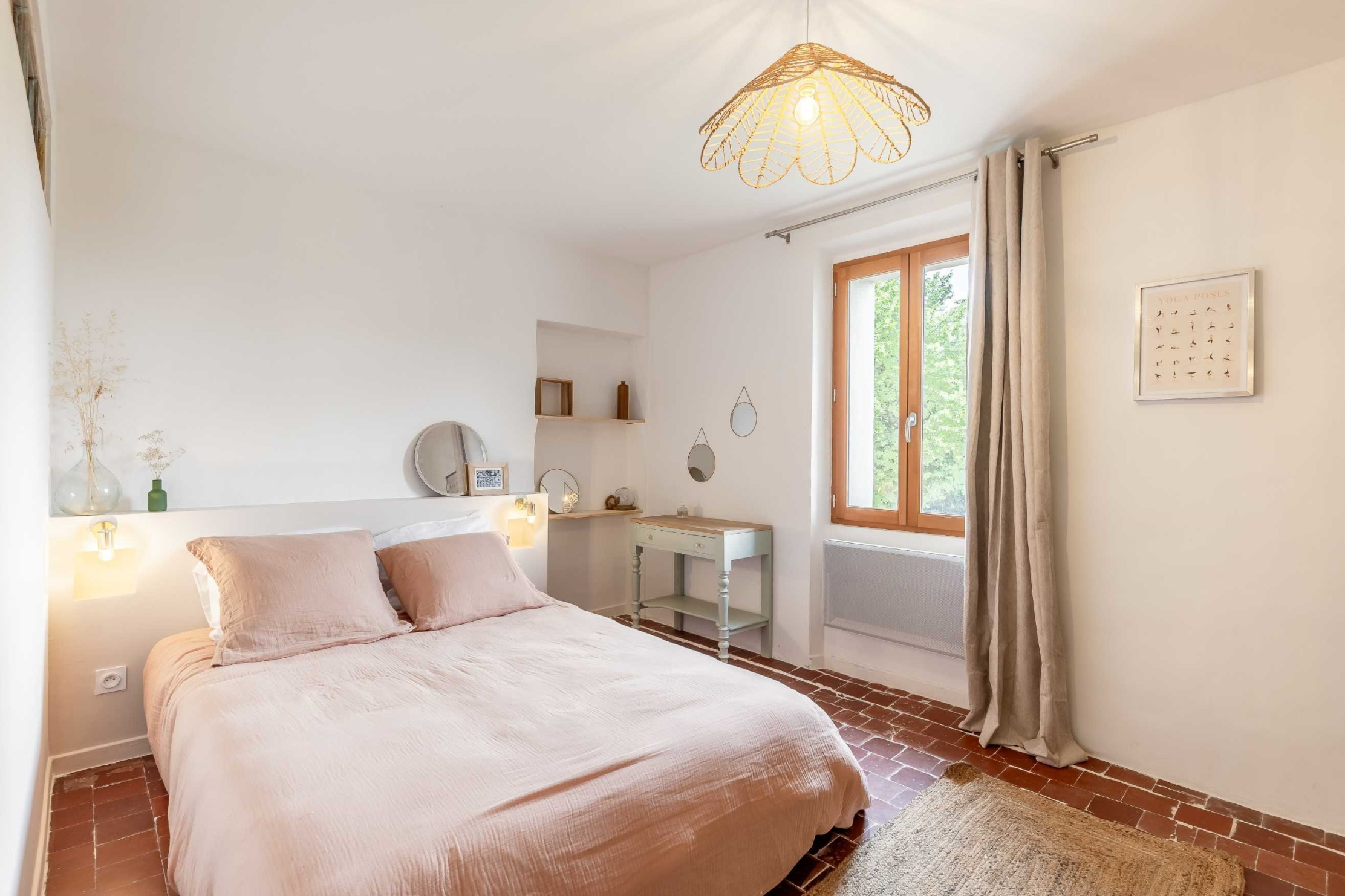 Une chambre à coucher lumineuse avec un lit double aux draps roses, des murs blancs, un sol en terre cuite, une fenêtre en bois, et une coiffeuse avec des miroirs.