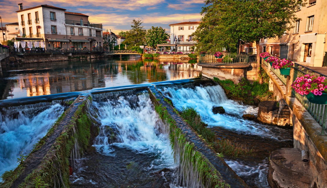 Une scène pittoresque de L'Isle-sur-la-Sorgue, avec une cascade en forme de V formée par la rivière bordée de murs moussus, encadrée par des bâtiments traditionnels, des terrasses de restaurant et des arbres verdoyants sous un ciel nuageux au coucher du soleil.