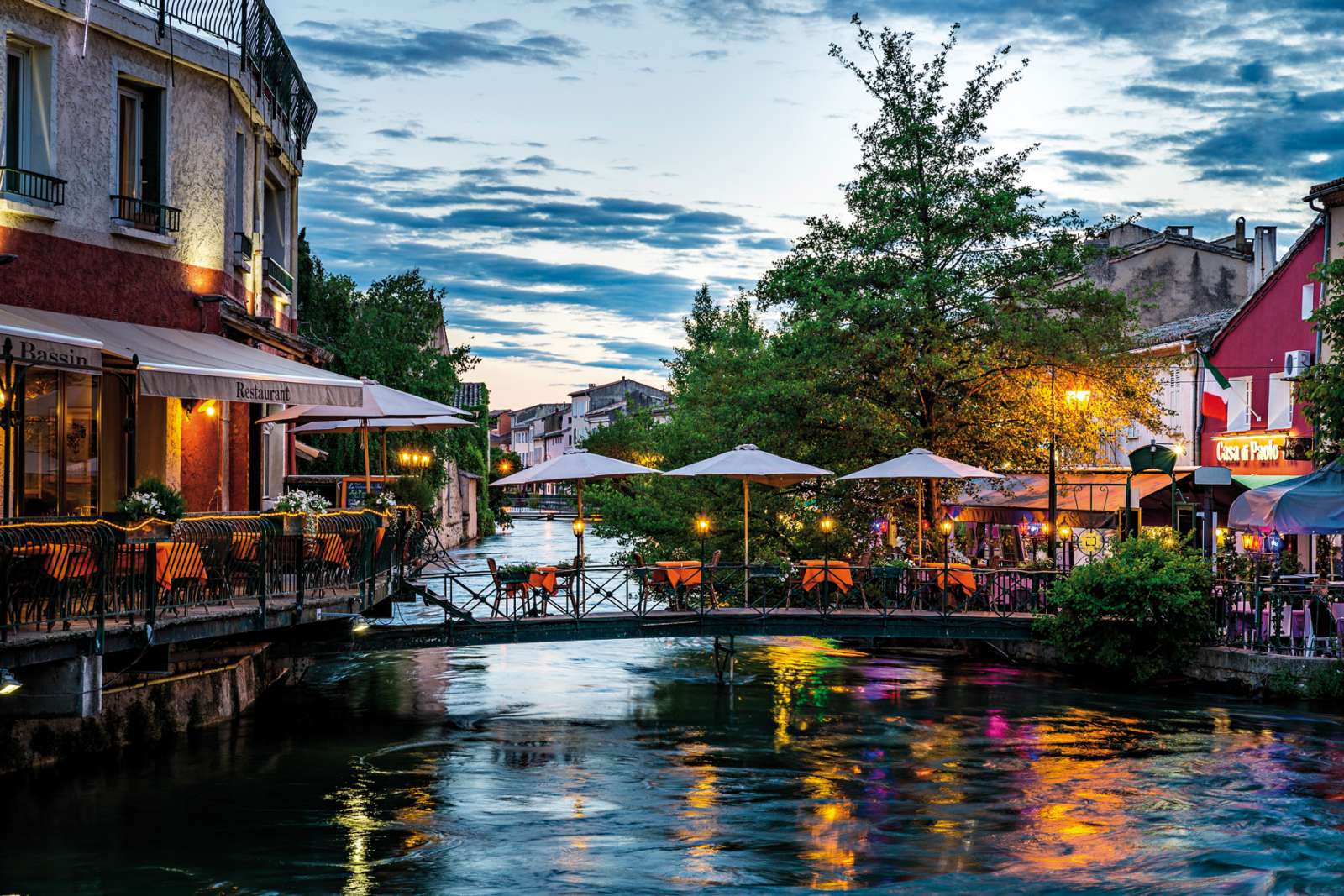 Une charmante vue nocturne de L'Isle-sur-la-Sorgue, avec des restaurants éclairés le long de la rivière, des tables sous des parasols, et un petit pont traversant l'eau.
