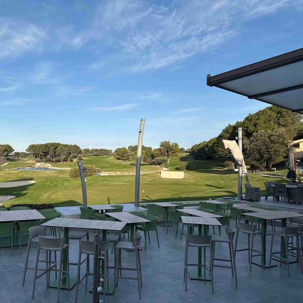 Une vaste terrasse extérieure avec de nombreuses tables et chaises vertes modernes, donnant sur un parcours de golf verdoyant sous un ciel bleu parsemé de nuages.