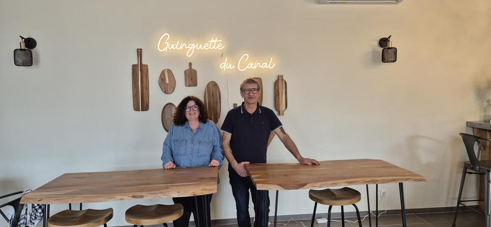 Deux personnes souriantes, une femme et un homme, se tiennent devant un mur beige avec une enseigne lumineuse "Guinguette du Canal" et des planches de bois décoratives, flanquées de deux tables en bois massif.