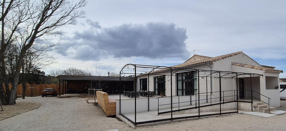 Un bâtiment clair de style provençal avec un toit en tuiles, flanqué d'une pergola en fer noir, d'une rampe d'accès, et d'une terrasse meublée, le tout sur un terrain de gravier sous un ciel nuageux, avec des arbres au fond.