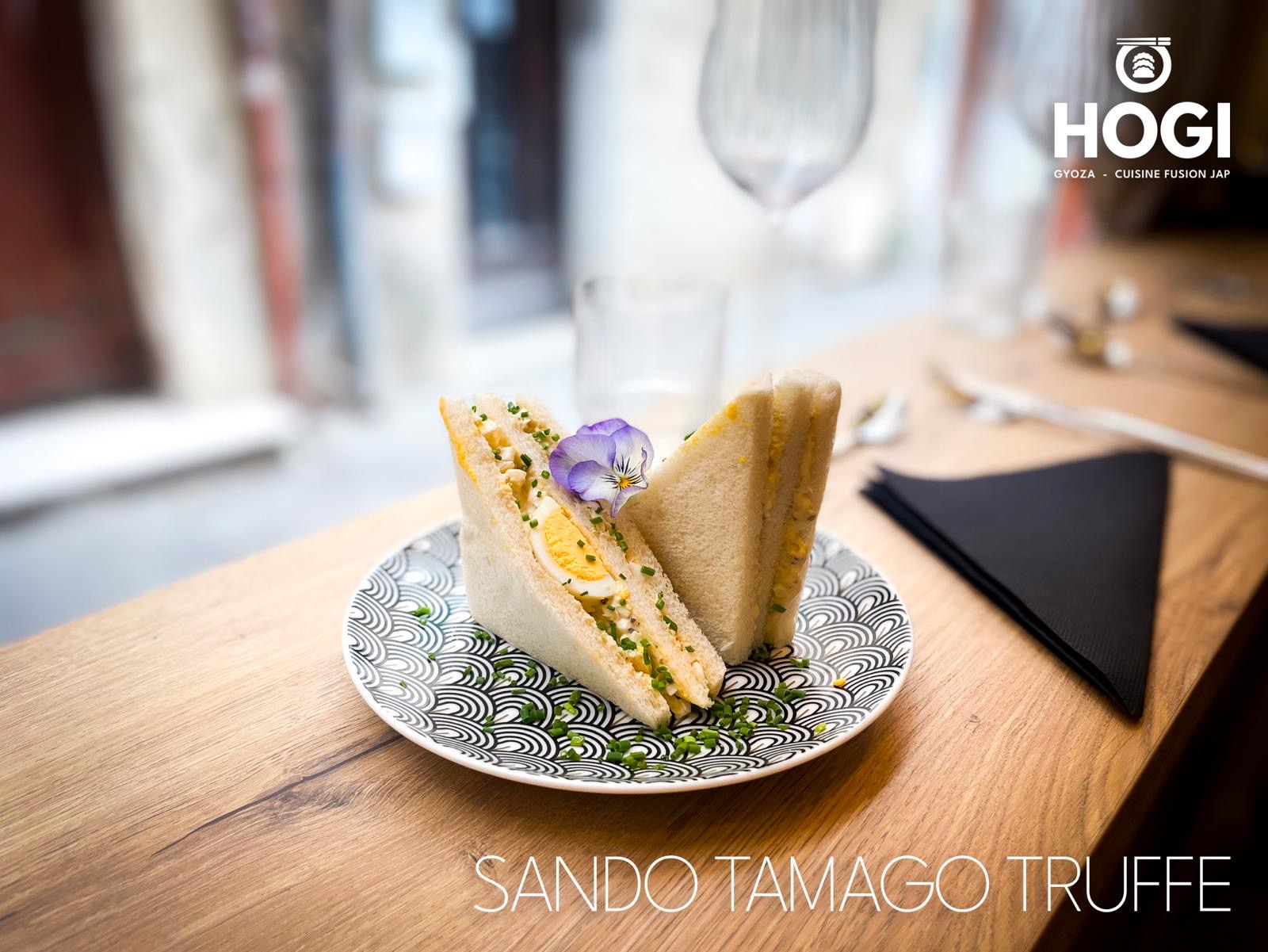 Un sandwich Sando Tamago Truffe garni d'œuf et d'une fleur comestible, coupé en triangles et servi sur une assiette à motifs noirs et blancs sur une table en bois.