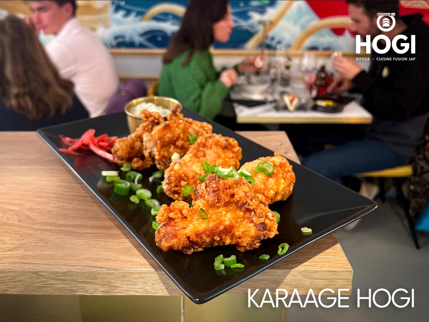 Un plat de Karaage Hogi, du poulet frit croustillant garni de ciboulette ciselée et accompagné de légumes marinés et d'une sauce blanche, est posé sur une table en bois dans un restaurant avec des clients en arrière-plan.