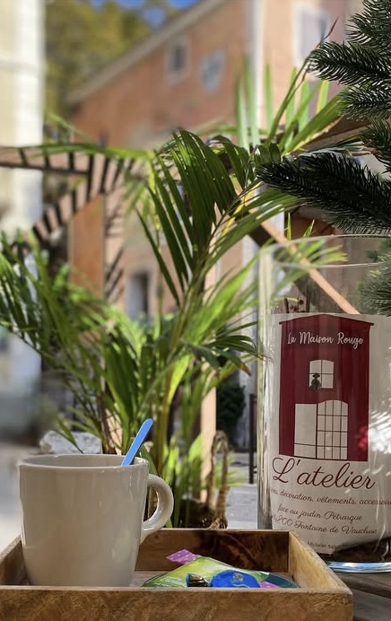 Une tasse blanche avec une cuillère bleue est posée sur un plateau en bois, à côté d'une bouteille en verre "La Maison Rouge L'atelier", avec des plantes vertes et un bâtiment flou en arrière-plan.