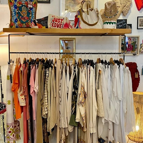 Une penderie remplie de vêtements de style bohème, principalement dans des tons clairs et neutres, est surmontée d'une étagère en bois présentant des accessoires comme des coussins, des sacs et des chapeaux.