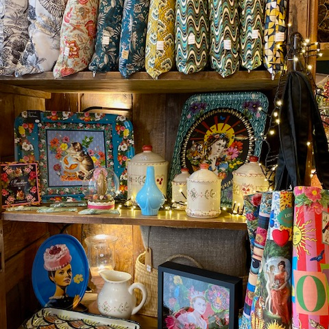 Une étagère en bois regorge d'objets décoratifs variés, incluant des coussins aux motifs colorés, des cadres avec des illustrations, des vases, des boîtes et des plats ornés de visages et de motifs floraux, créant une ambiance bohème et éclectique.