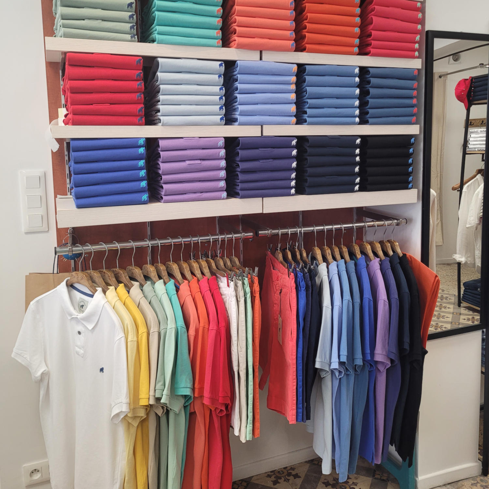 Un étalage de vêtements, avec des piles de chemises polo colorées sur des étagères au-dessus d'une rangée de cintres présentant des t-shirts et pantalons de différentes couleurs.