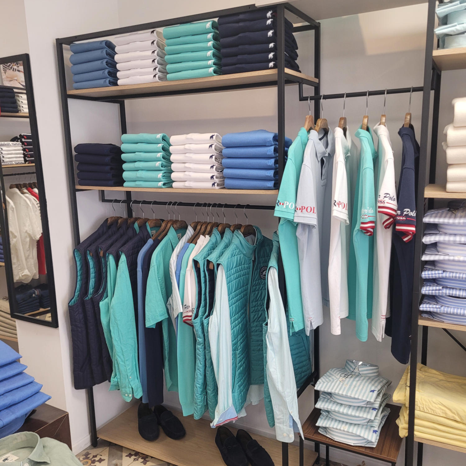 Un magasin de vêtements avec des étagères en bois et métal remplies de piles de polos pliés dans des tons de bleu, blanc et turquoise, et des vêtements suspendus sur des cintres, incluant des polos, des gilets et des chemises.