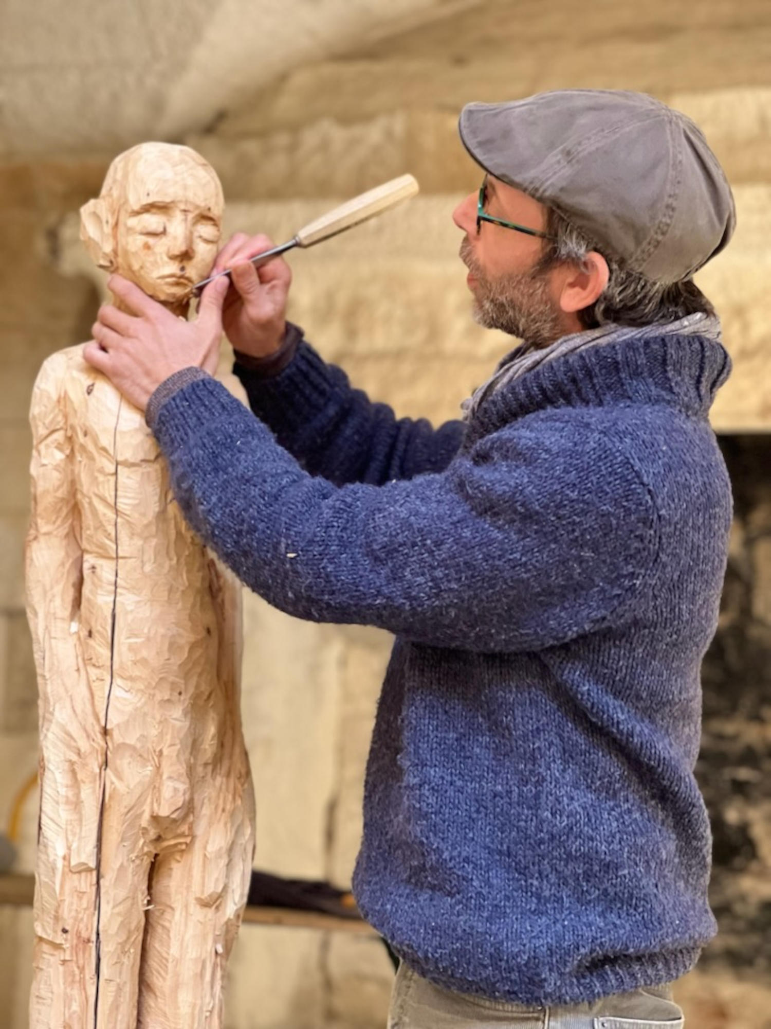 Un sculpteur barbu avec une casquette et des lunettes, travaillant méticuleusement sur une sculpture en bois d'une silhouette humaine, utilisant un ciseau.