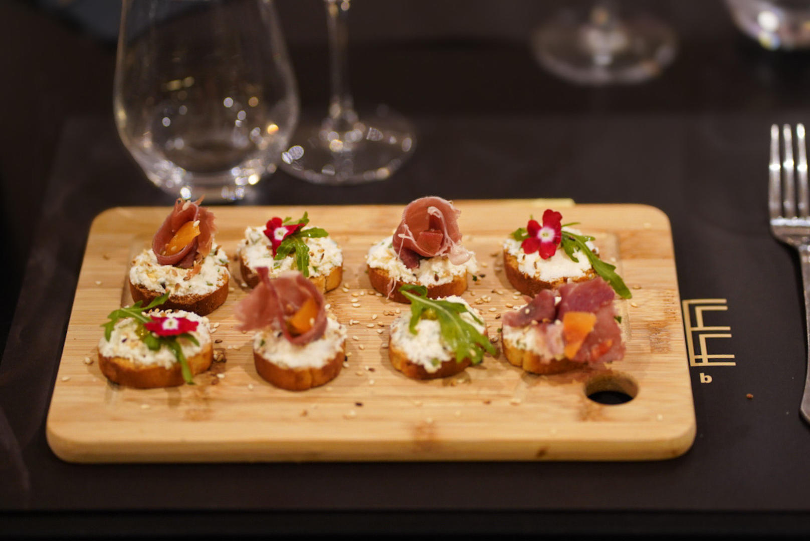 Huit petites bruschettas garnies de fromage frais, de charcuterie ou de poisson, et de verdure, sont présentées sur une planche en bois, avec des verres en arrière-plan.