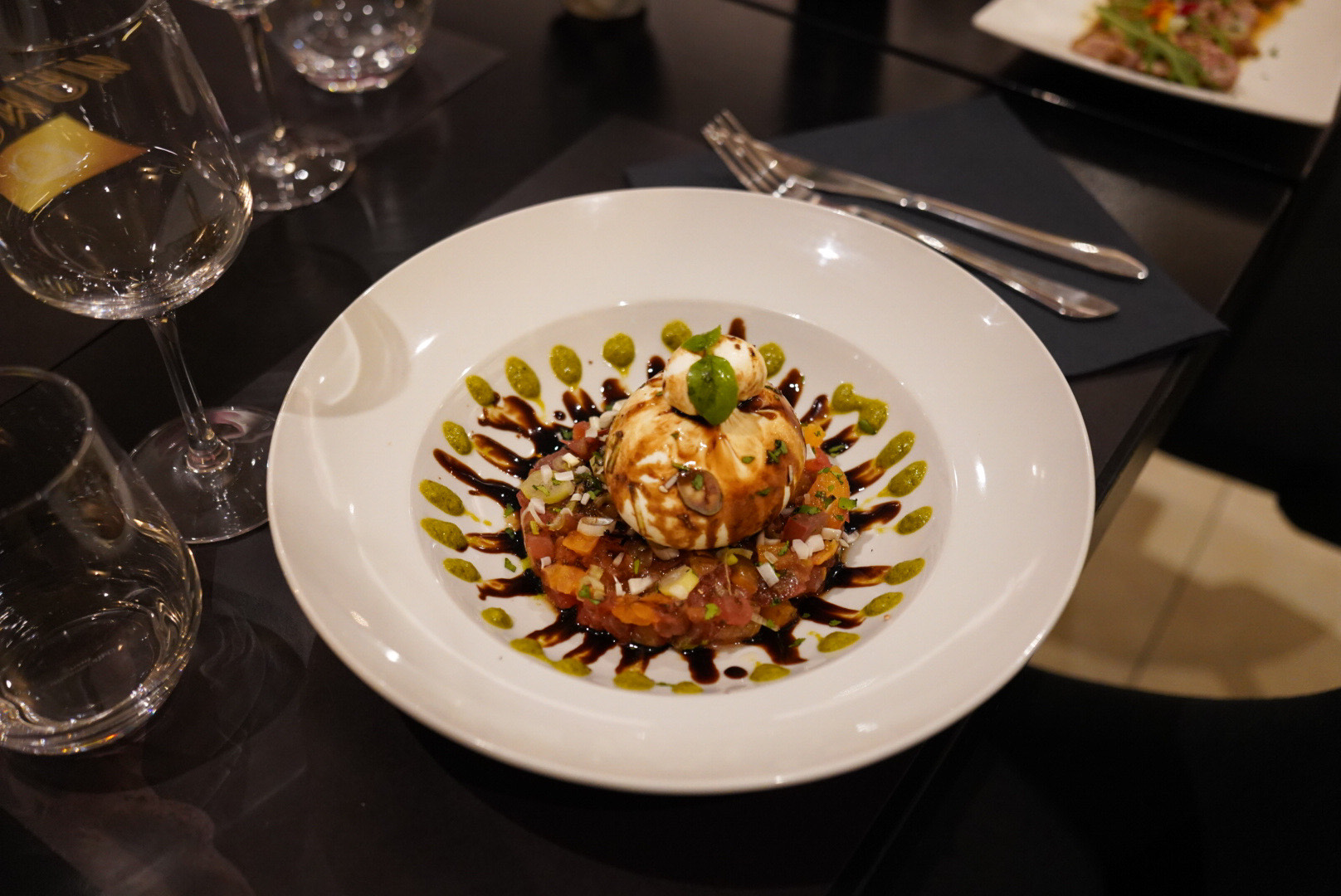 Un plat gastronomique avec un dôme de nourriture garni de basilic frais, reposant sur une base de légumes coupés en dés et de sauce au vinaigre balsamique, avec une garniture verte pointillée sur le bord de l'assiette blanche, et des verres à vin en arrière-plan.