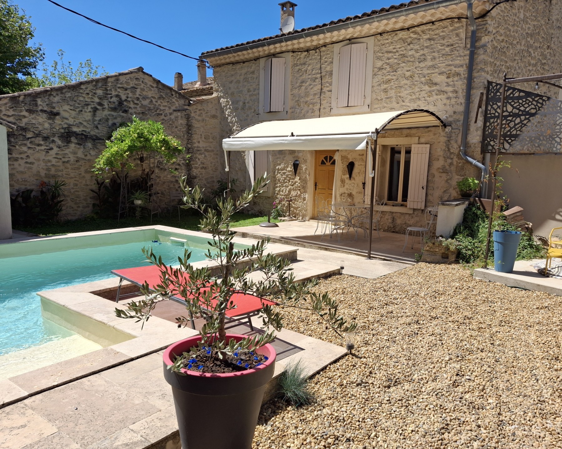 Une villa provençale en pierre avec une piscine, une terrasse en gravier aménagée avec une pergola et une zone repas, et un olivier en pot au premier plan.