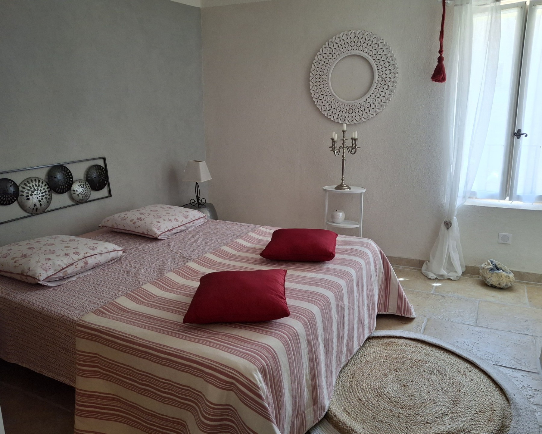 Une chambre lumineuse avec un lit double orné d'un couvre-lit rayé rouge et blanc et de coussins rouges, une tête de lit en métal avec des sphères décoratives, une table de chevet avec une lampe, et une grande fenêtre à rideaux blancs.