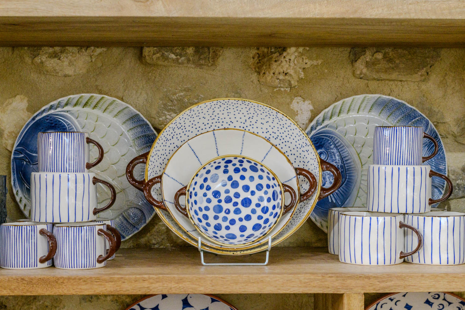 Un ensemble de vaisselle aux motifs bleus et blancs, incluant des assiettes, des bols et des tasses, est exposé sur une étagère en bois avec un mur de pierre en arrière-plan.