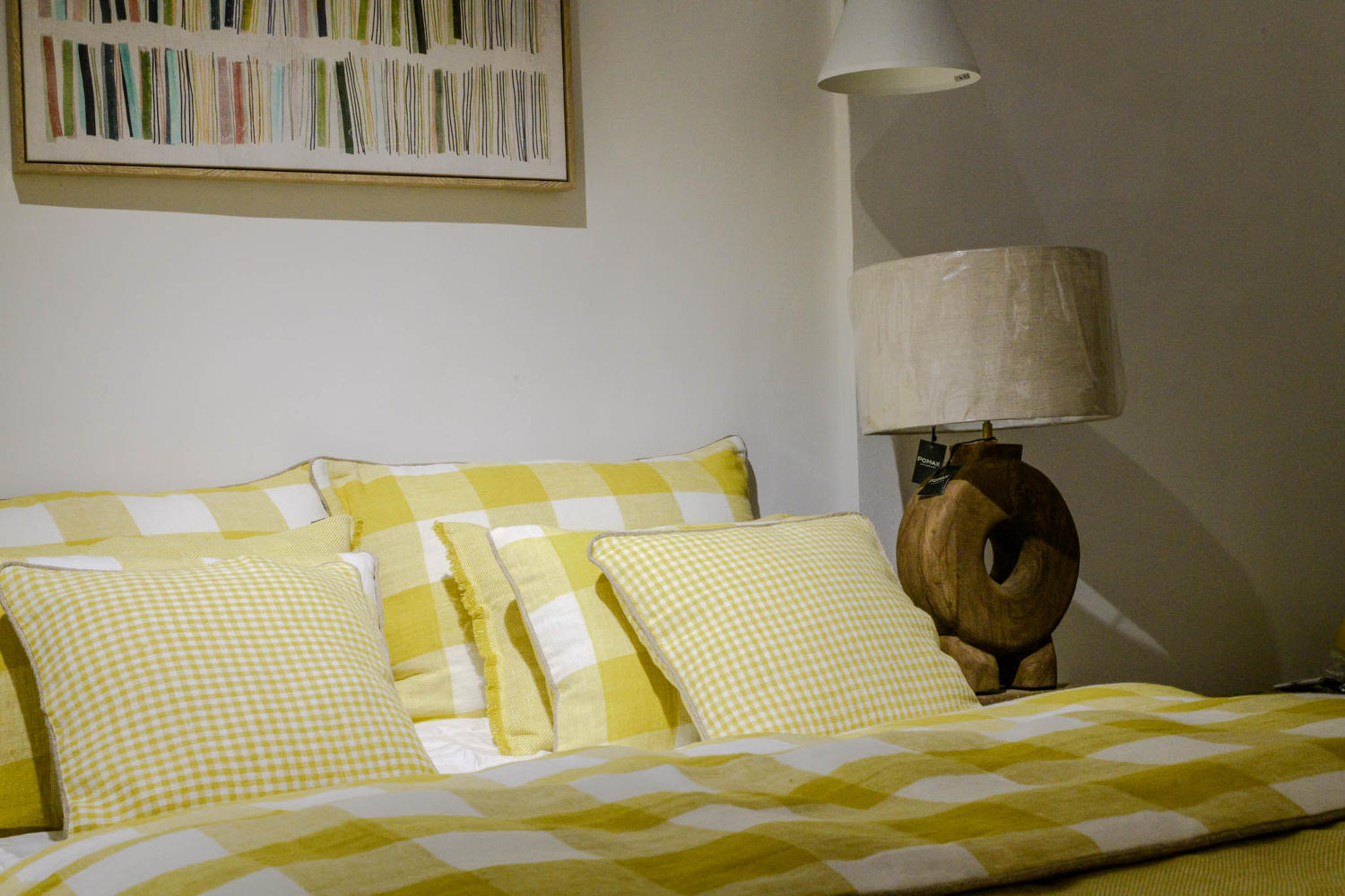 Une chambre à coucher présente un lit double avec une literie à carreaux jaunes et blancs, des coussins assortis, une lampe de chevet en bois et une peinture abstraite aux couleurs vives au-dessus du lit.