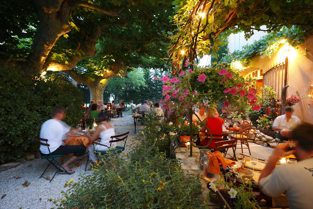Une vaste terrasse extérieure de restaurant sous d'imposants platanes feuillus, avec des tables dressées pour les convives, des parasols et des guirlandes lumineuses créant une ambiance chaleureuse en fin de journée.