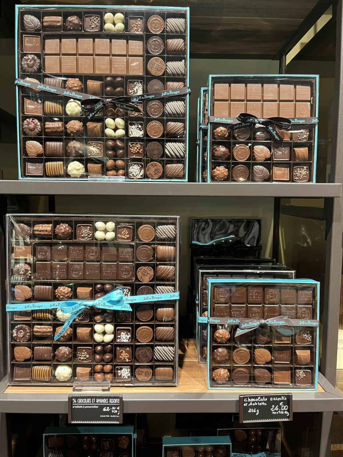 De grandes boîtes de chocolats assortis, certains attachés avec des rubans bleus, exposées sur des étagères dans un magasin.