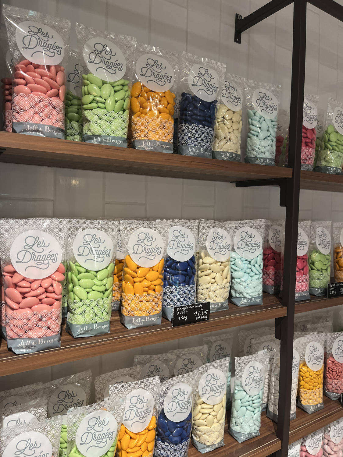 Des sachets de dragées colorées, de différentes teintes pastel, disposés sur des étagères en bois dans un magasin.