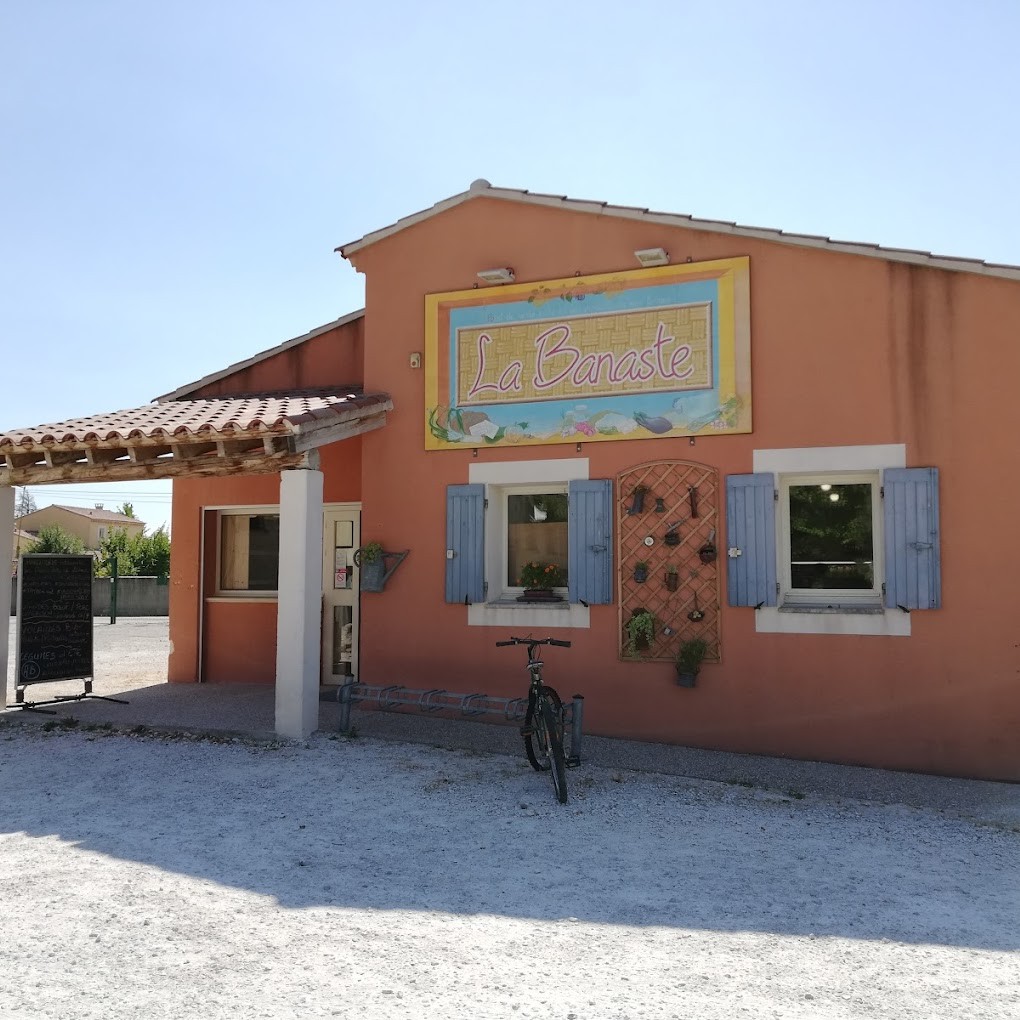 Un bâtiment de couleur pêche avec un grand panneau "La Banaste" au-dessus de deux fenêtres à volets bleus. Une bicyclette est garée devant l'entrée ombragée par un auvent, avec un panneau de menu sur le côté.
