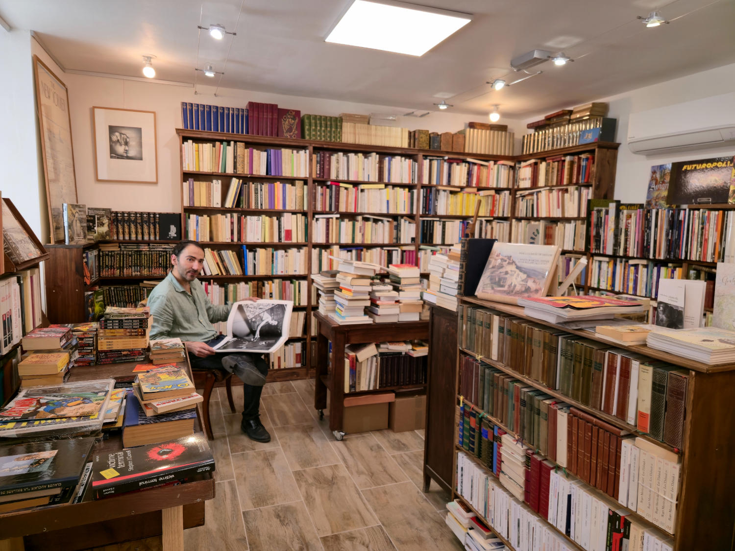 Un homme barbu est assis dans une librairie bien fournie, entouré d'étagères du sol au plafond remplies de livres. Des piles de livres sont également empilées sur une table en bois et par terre.