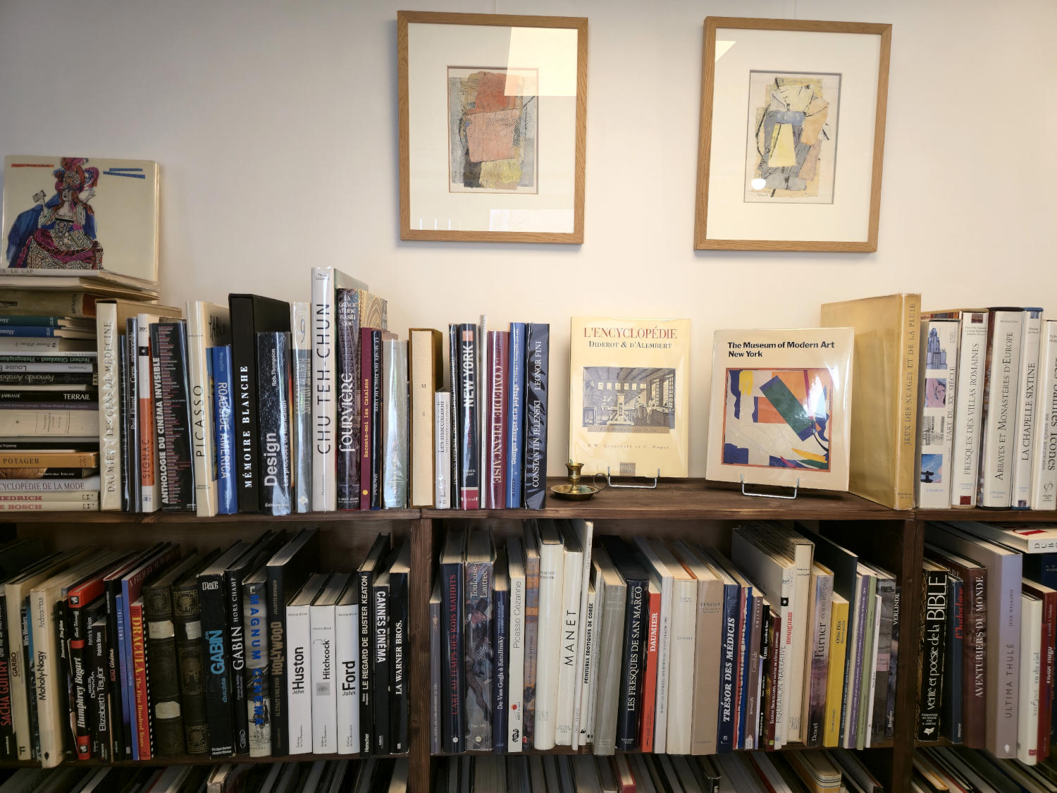 Une bibliothèque en bois remplie de livres aux reliures variées, avec deux cadres d'œuvres d'art abstraites accrochés au mur blanc au-dessus.