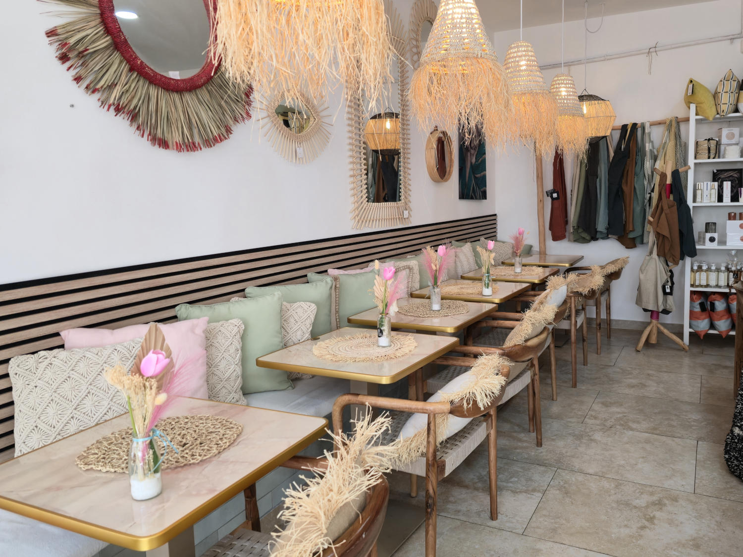 Un café-boutique lumineux et moderne avec une rangée de tables en marbre bordant une banquette garnie de coussins roses et verts. Le décor est bohème avec des luminaires, miroirs et décorations murales en fibres naturelles.