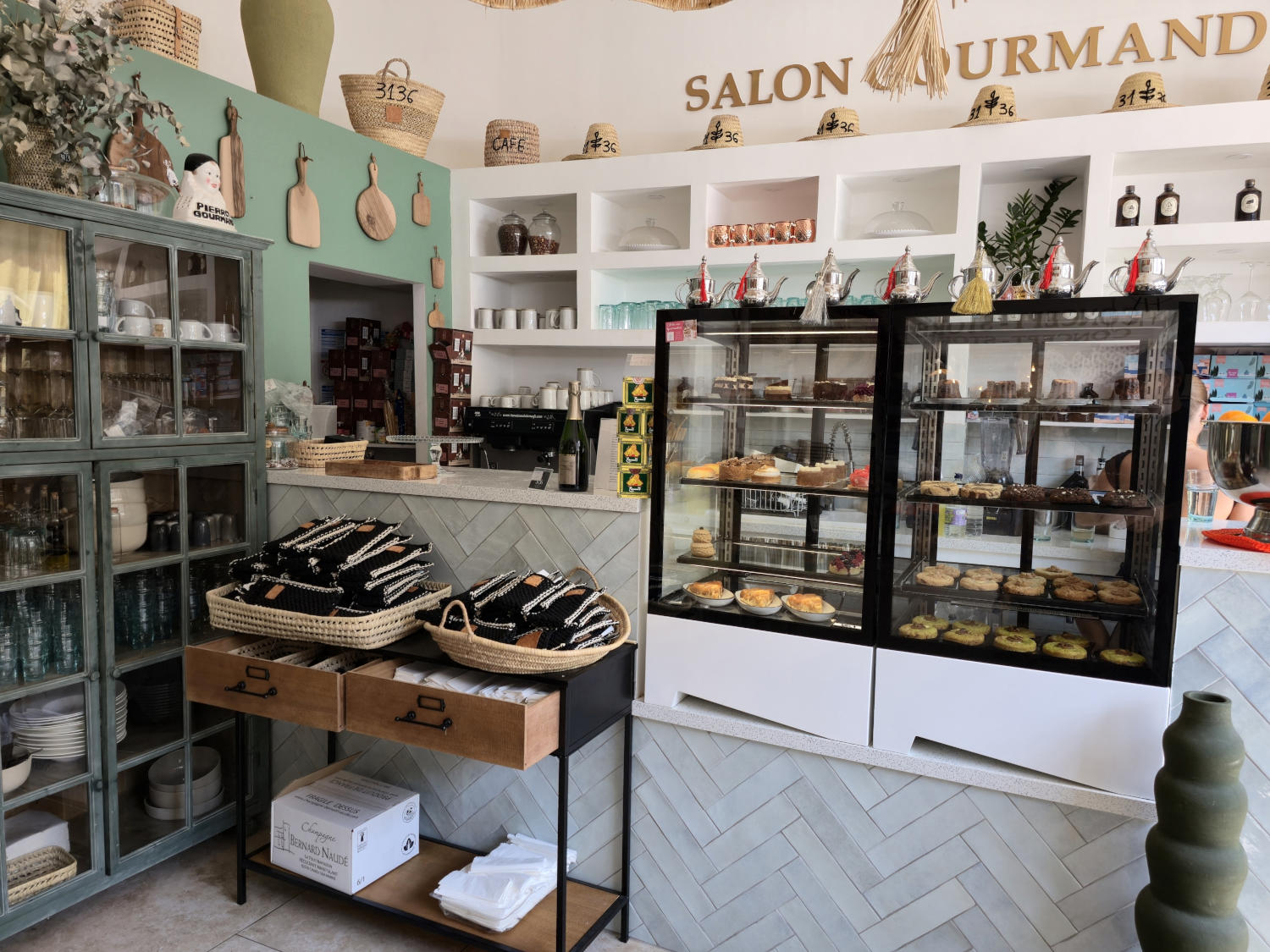 Un café lumineux et moderne avec des étagères blanches remplies de vaisselle et d'articles de décoration, et une vitrine réfrigérée présentant des pâtisseries. Des paniers de produits emballés sont exposés au premier plan, et les murs sont décorés de tons pastel.