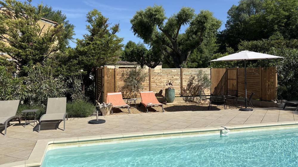 **Photo 3 – L’espace piscine et détente** Cette photo présente un agréable espace extérieur aménagé autour d’une piscine, dans un cadre verdoyant et paisible. * Au premier plan, une piscine aux eaux claires et turquoise occupe une grande partie de l’image. * Une vaste terrasse carrelée entoure le bassin, offrant un espace confortable pour se détendre. * Plusieurs bains de soleil sont disposés autour de la piscine, certains dans des tons gris et d’autres avec des coussins corail qui apportent une touche de couleur. * Un grand parasol blanc crée une zone ombragée à proximité d’un petit salon de jardin composé de fauteuils et d’une table. * Des claustras en bois délimitent l’espace et préservent l’intimité des lieux tout en s’intégrant harmonieusement à l’environnement. * Une grande jarre décorative et des plantations ornementales ajoutent du charme à l’aménagement. * De nombreux arbres et arbustes entourent la propriété, créant une atmosphère calme et naturelle. * Le ciel bleu et l’ensoleillement généreux renforcent l’impression de vacances et de bien-être. L’ensemble évoque un espace de détente élégant et convivial, idéal pour profiter des journées ensoleillées dans un environnement préservé.