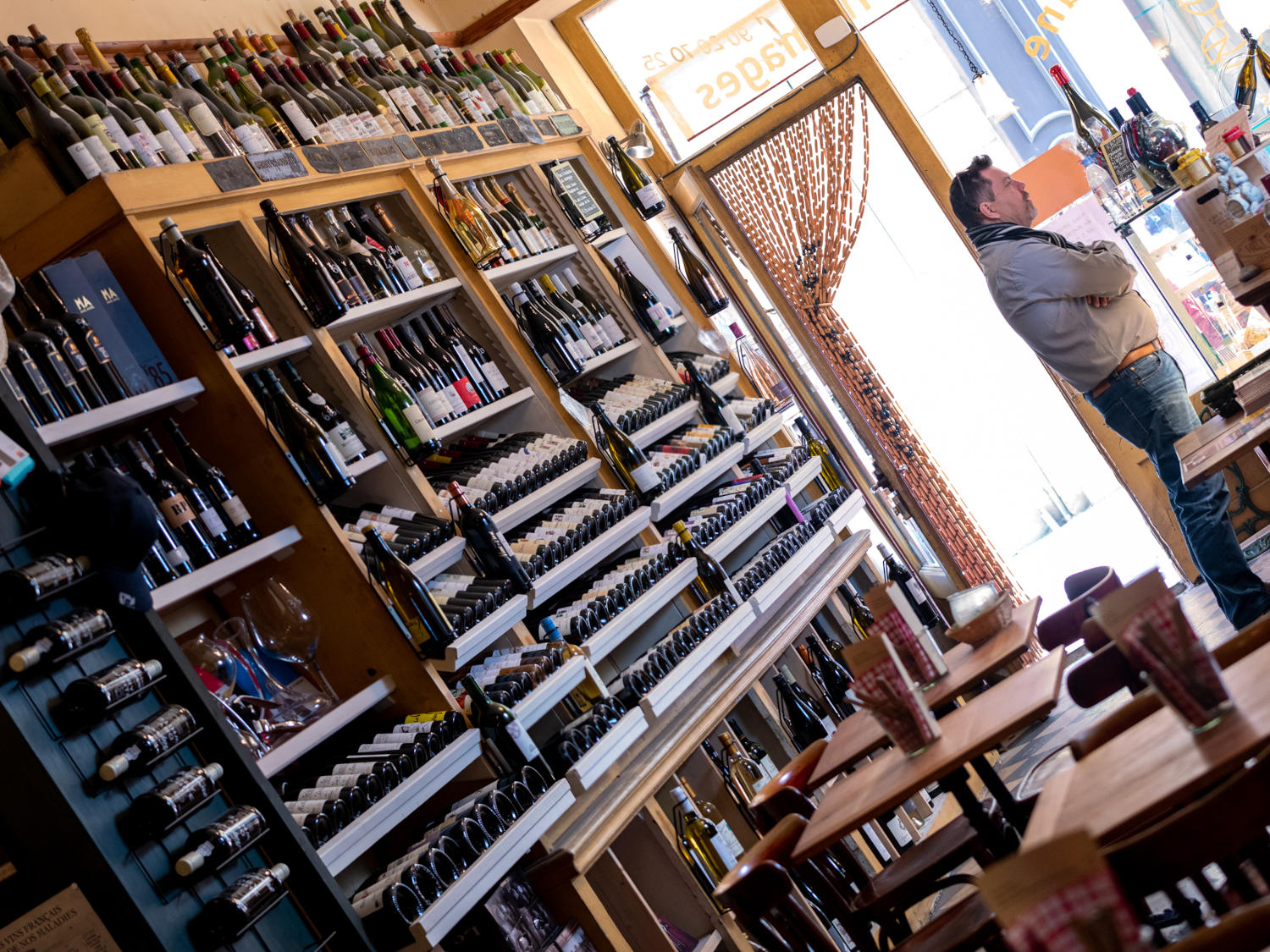 L'intérieur d'une cave à vins ou d'un magasin de spiritueux, avec des étagères remplies de bouteilles de vin s'étendant du sol au plafond. Un homme se tient près de l'entrée, éclairé par la lumière du jour.