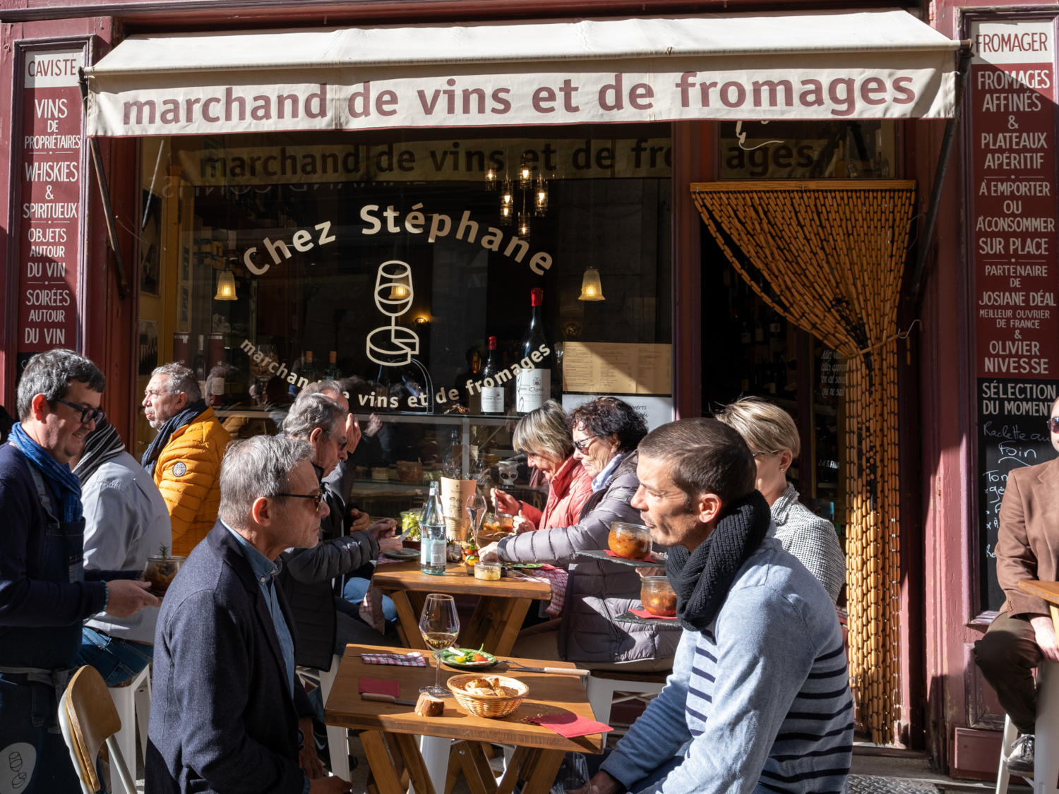 Une foule de personnes est attablée à l'extérieur d'un restaurant "Marchand de vins et de fromages Chez Stéphane". Elles dégustent des boissons et de la nourriture, tandis que d'autres s'approchent de la façade vitrée.