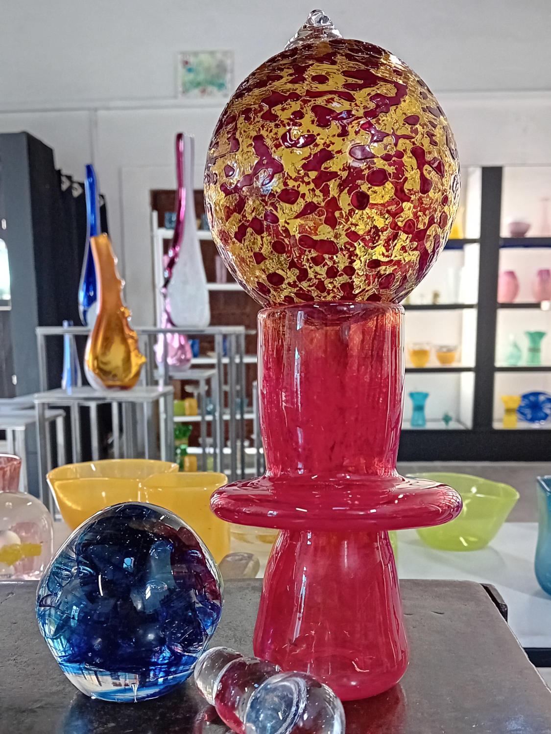  Pièces d’art verrier colorées exposées en gros plan dans un atelier : une grande sculpture en verre soufflé composée d’une base rouge et violette surmontée d’une sphère transparente aux reflets roses, accompagnée d’autres objets décoratifs. 