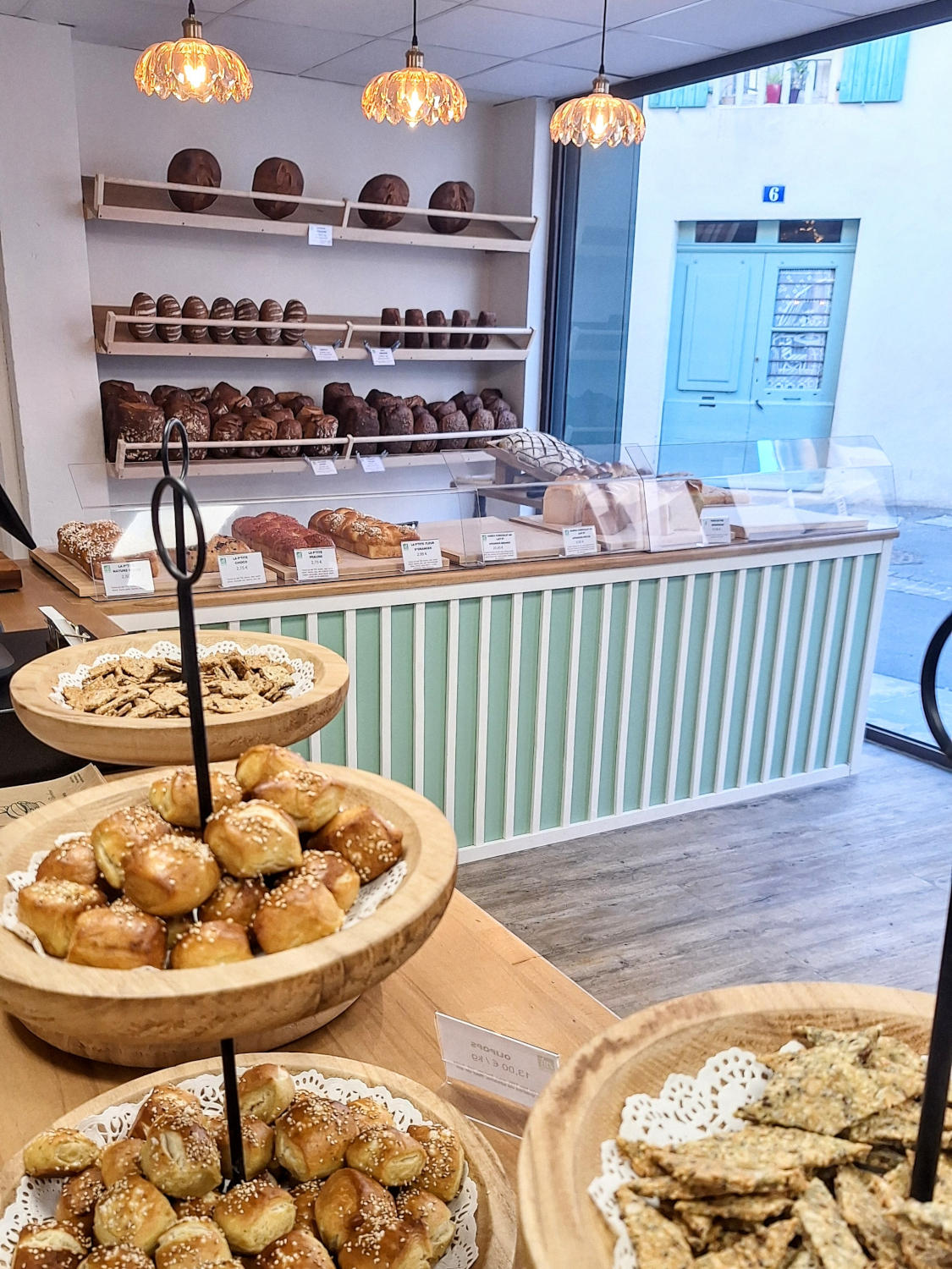 L'intérieur d'une boulangerie moderne et lumineuse, avec un comptoir vert menthe et blanc, derrière lequel sont exposés divers pains. Au premier plan, des bols en bois sont remplis de petits pains et de biscuits salés.