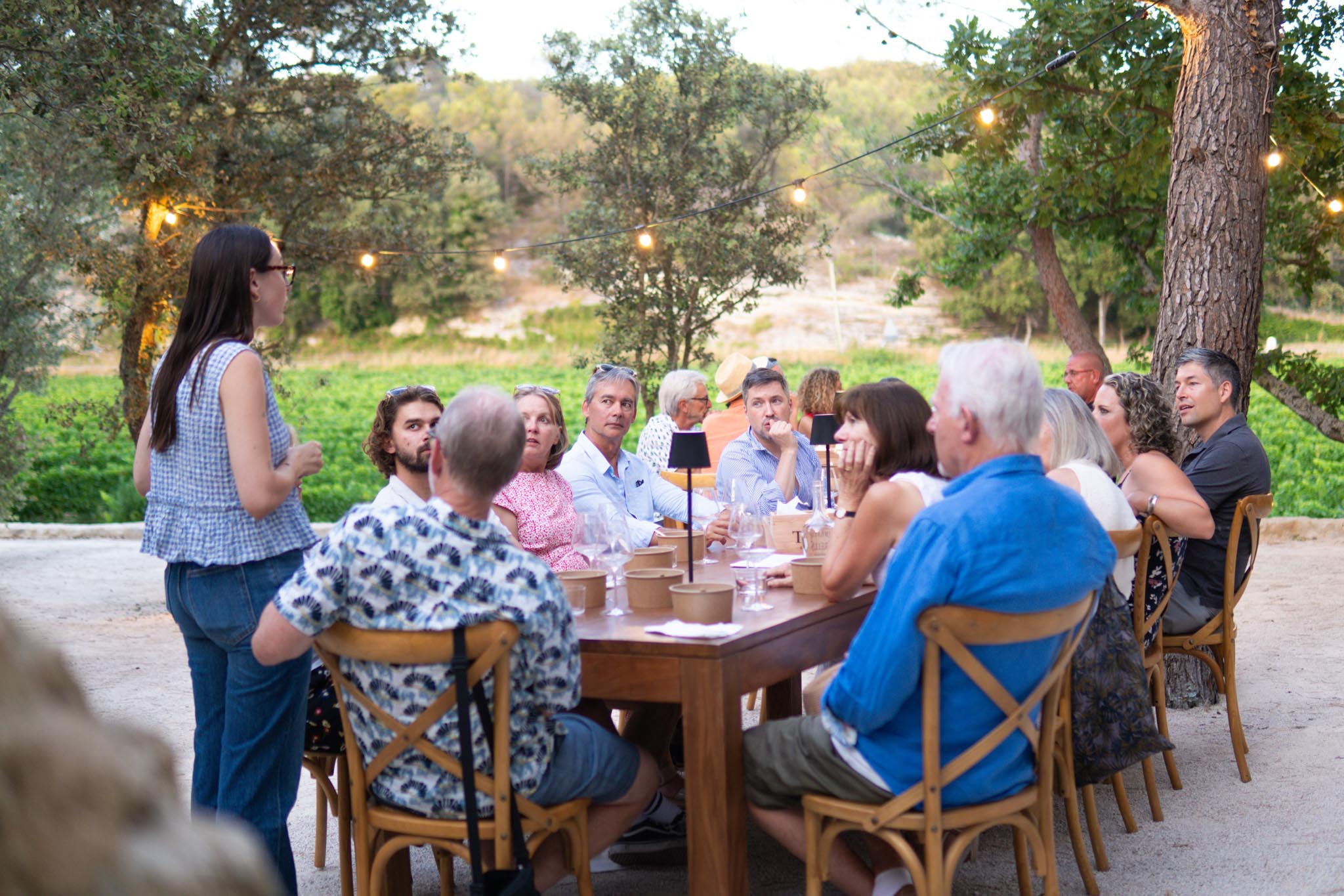 Un groupe de personnes est assis autour d'une longue table en bois pour un dîner en plein air, dans un cadre verdoyant. Une jeune femme se tient debout à l'extrémité de la table et s'adresse au groupe. Des guirlandes lumineuses sont suspendues au-dessus d'eux.