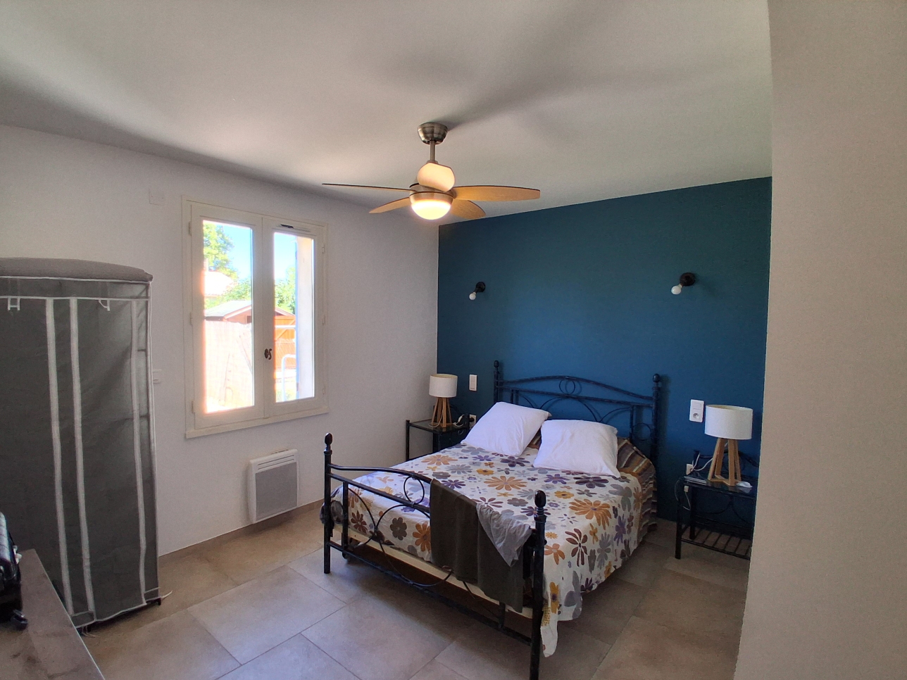 Une chambre lumineuse et bien éclairée avec un mur d'accent bleu marine derrière un lit double en fer forgé, paré d'une couette à motifs floraux et de deux oreillers blancs. Un ventilateur de plafond et deux tables de chevet avec des lampes sont également visibles.
