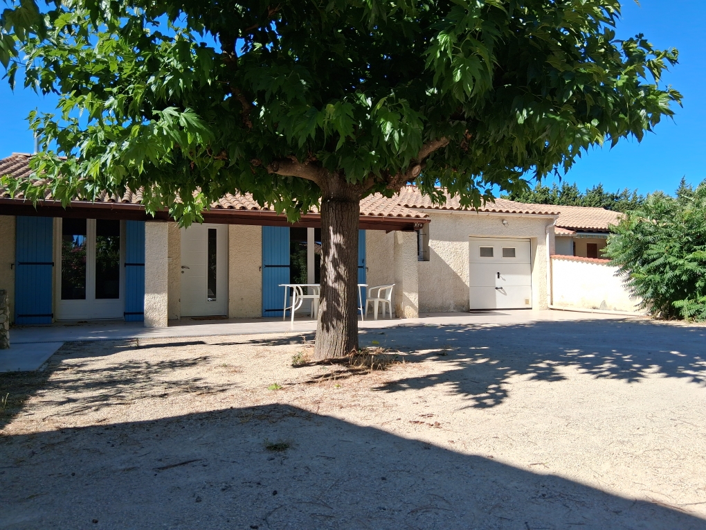 Une maison provençale à un étage, avec des volets bleus et un garage blanc, sous un grand arbre feuillu offrant de l'ombre sur la terrasse et l'allée en gravier. Le ciel est clair et bleu.
