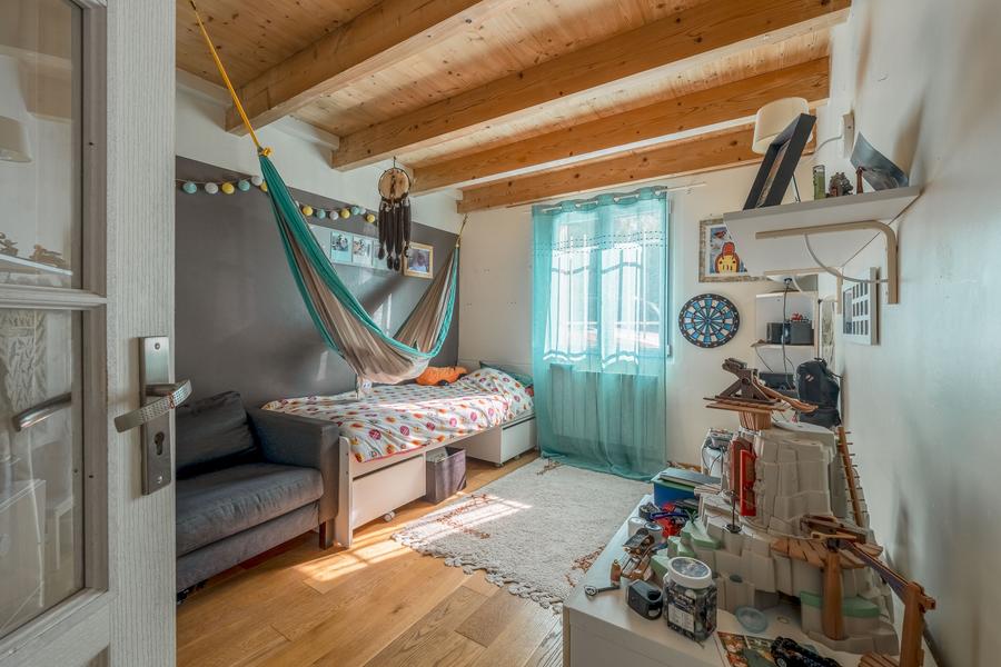 Une chambre d'enfant avec des poutres en bois apparentes au plafond. Elle est meublée d'un lit simple avec une literie colorée, d'une petite table de chevet avec des jouets dessus, d'un canapé gris et d'un hamac suspendu au-dessus du lit. Une fenêtre avec des rideaux bleus laisse entrer la lumière du soleil.