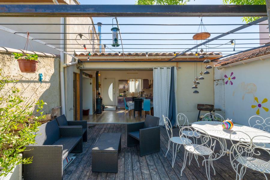 Une terrasse extérieure avec un plancher en bois, meublée d'un canapé gris, de fauteuils assortis et d'une table à manger blanche avec des chaises. Le patio est partiellement couvert par une pergola décorée de guirlandes lumineuses, de mangeoires à oiseaux et de plantes suspendues. Une porte ouverte révèle l'intérieur d'une maison à l'arrière-plan.