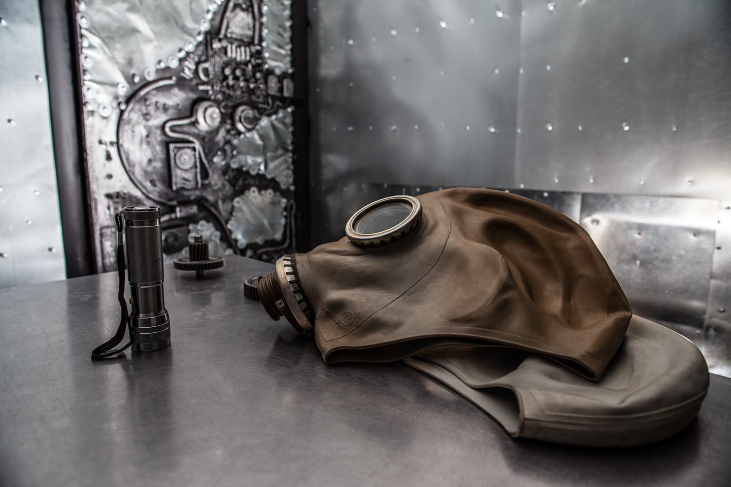 Un masque à gaz vintage beige et une lampe de poche en métal sont posés sur une table de métal, devant un mur texturé d'inspiration industrielle ou science-fiction.