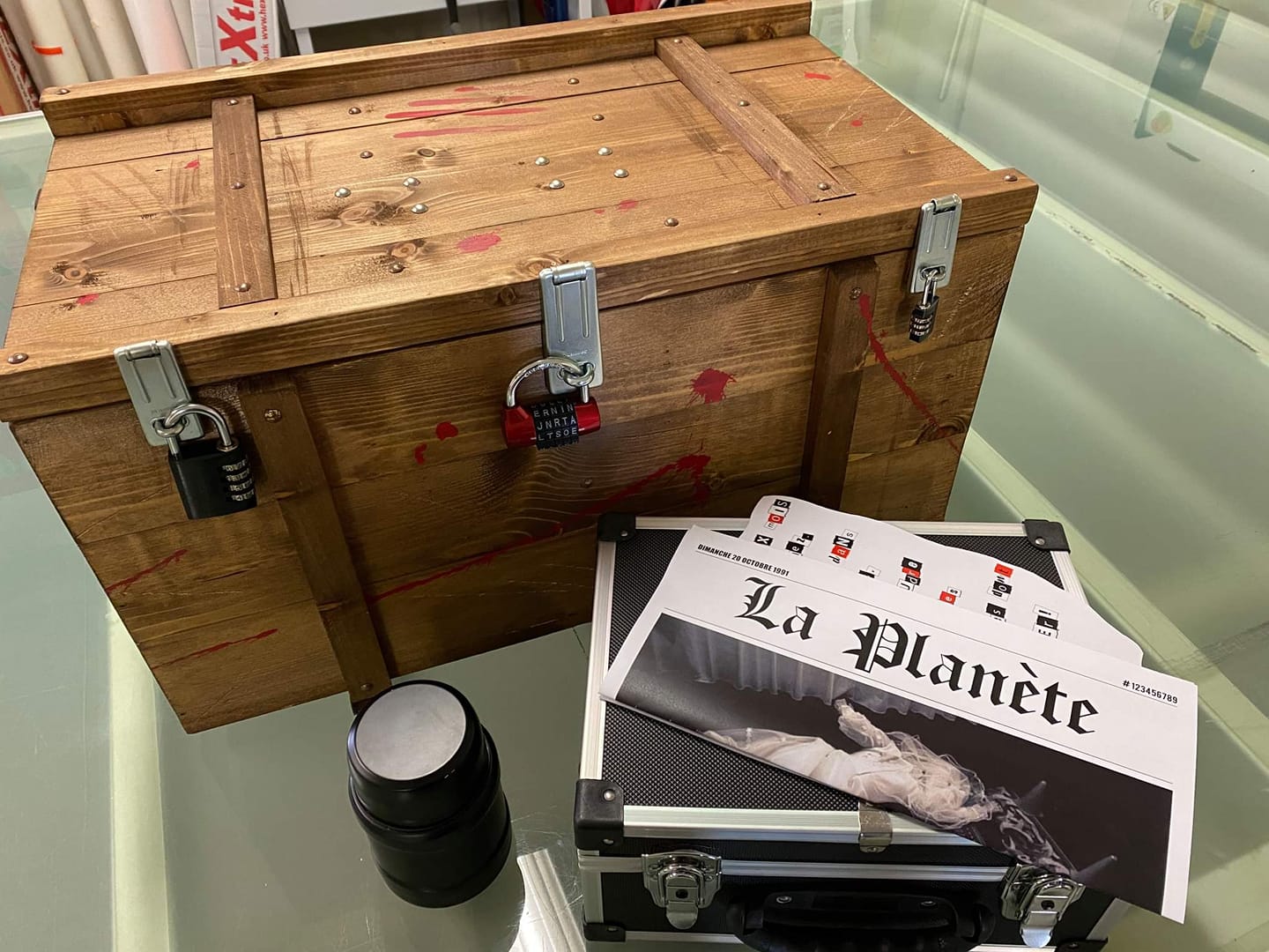 On voit une grande caisse en bois fermée par trois cadenas. Elle porte plusieurs traces rouges qui évoquent du sang. Devant la caisse se trouvent une mallette noire, un récipient cylindrique et des feuilles intitulées « La Planète ». L’ensemble donne l’impression d’une scène d’enquête ou d’un jeu d’énigmes.