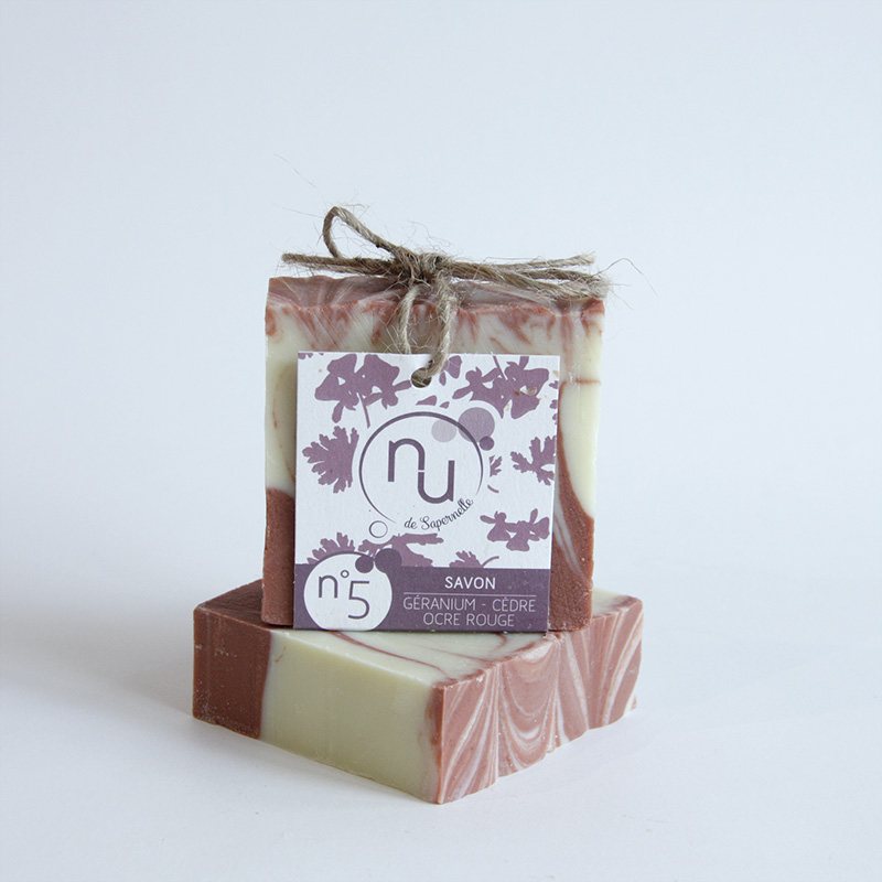 Deux savons artisanaux marbrés de crème et de rose-brun, surmontés d'une étiquette attachée avec de la ficelle, indiquant "Savon Géranium - Cèdre Ocre Rouge" et le logo "Nu de Sapernelle".