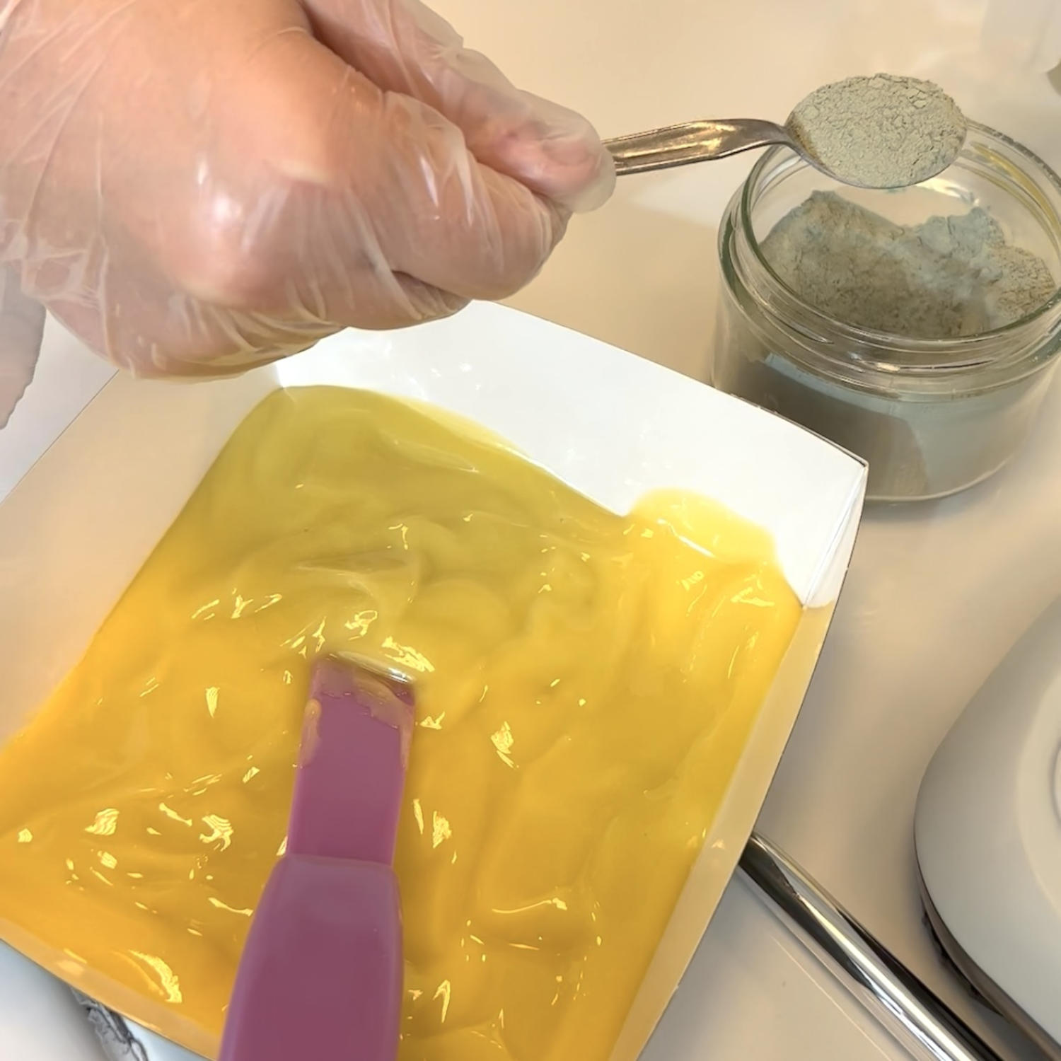 Une personne gantée verse une poudre gris-vert d'une cuillère dans un récipient blanc contenant une préparation jaune à l'aide d'une spatule mauve.