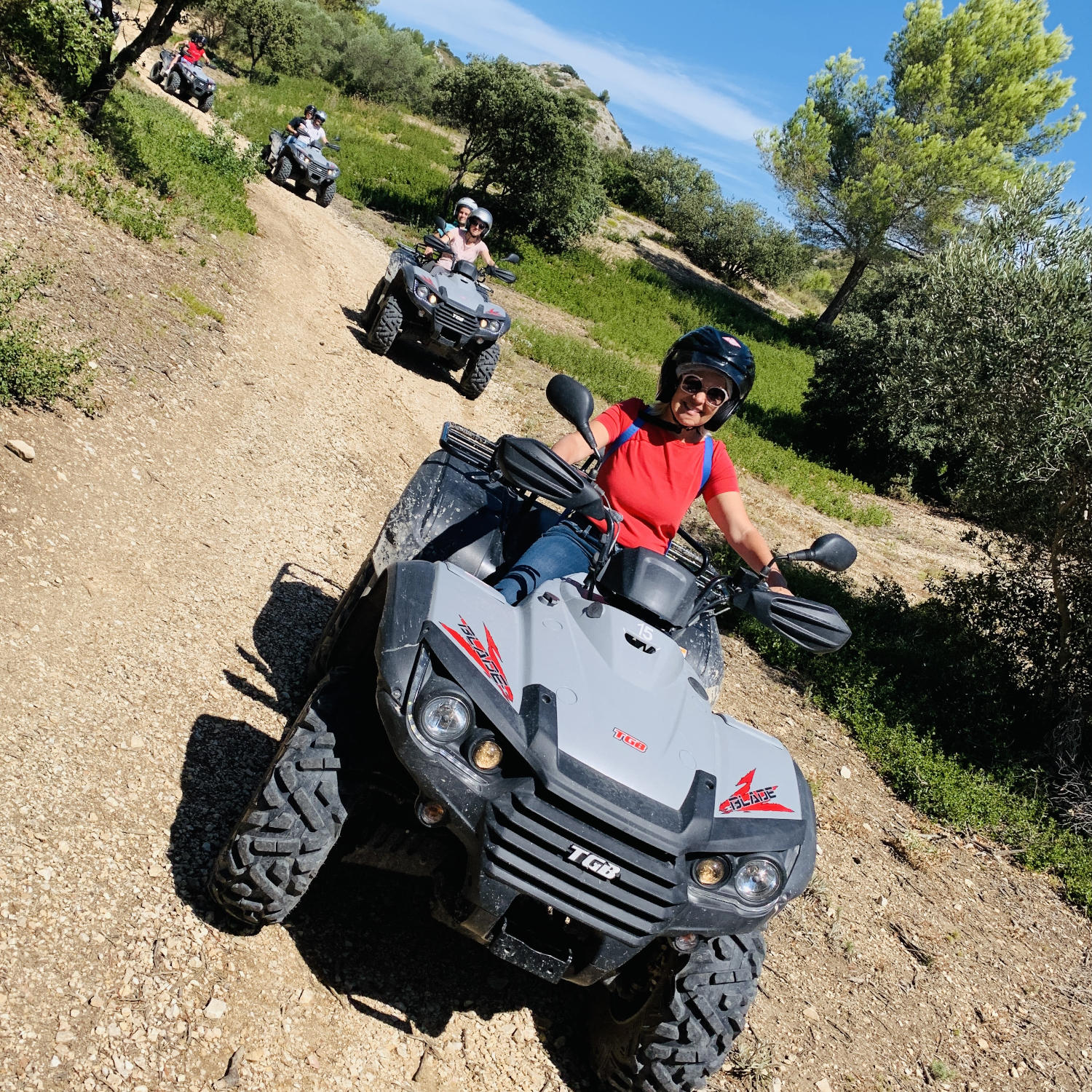 Un groupe de personnes sur des quads, roulant sur un chemin de terre à la campagne sous un ciel bleu.