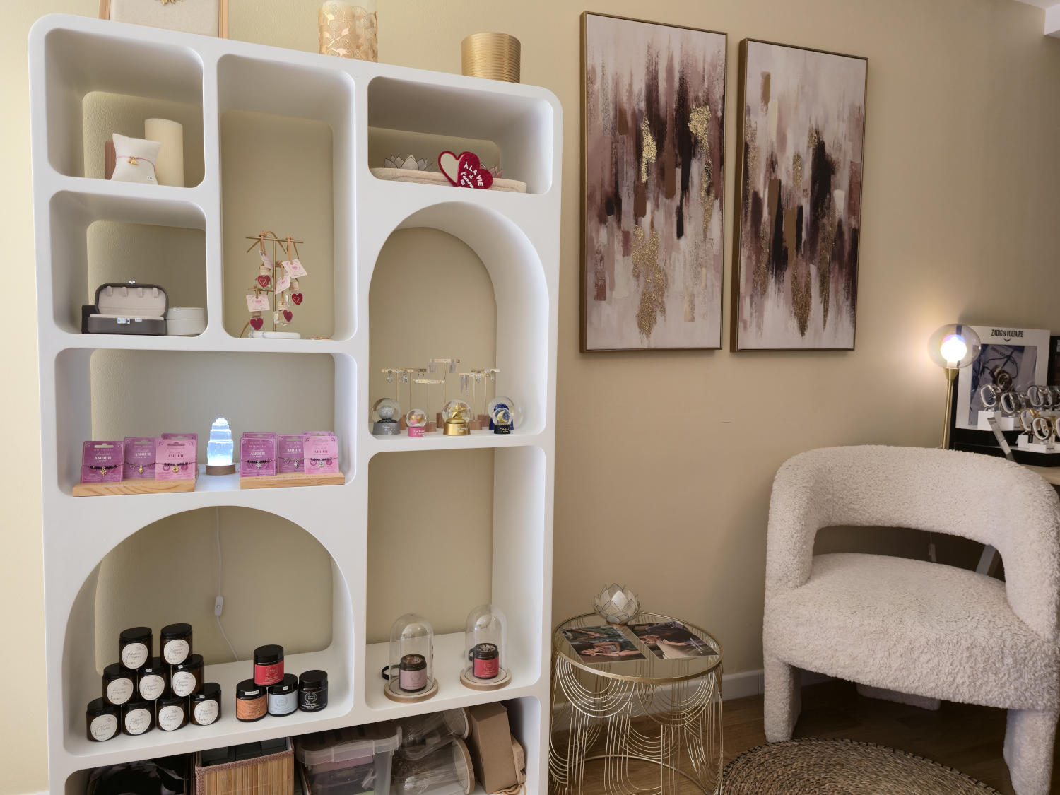 Un meuble de rangement blanc à arcades, rempli de produits de beauté, de décorations et d'articles de bien-être, à côté d'un fauteuil bouclette blanc et de tableaux abstraits.
