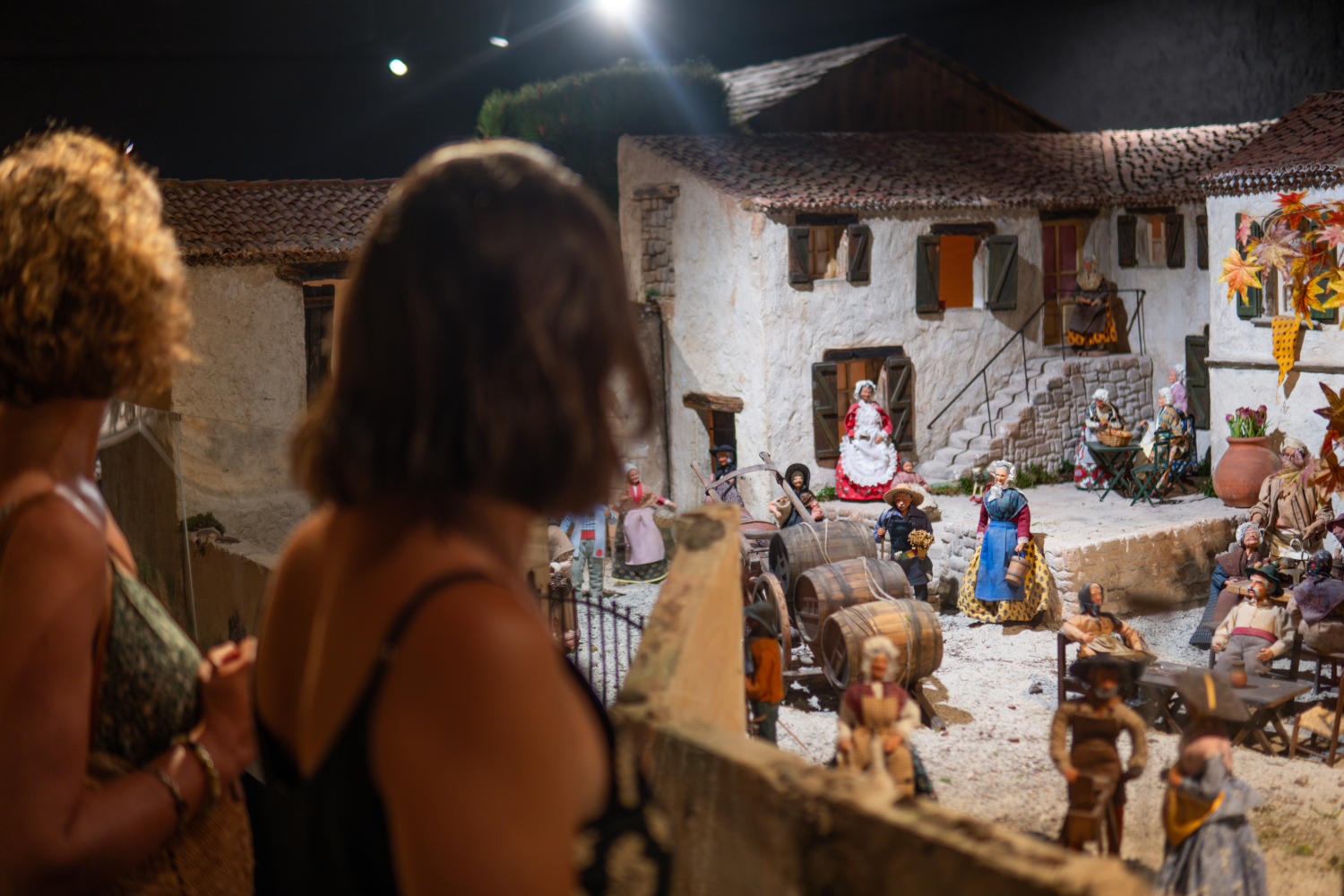 Deux femmes observent de dos une maquette de village provençal avec des figurines, représentant une scène de vie quotidienne.