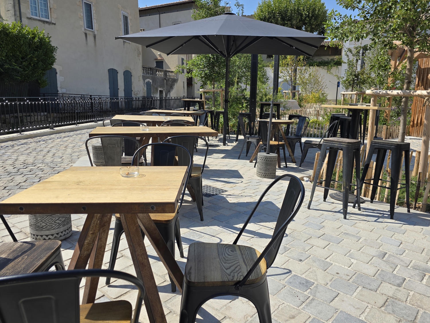 Une terrasse de café avec des tables et des chaises en bois et en métal, un grand parasol gris et un sol pavé.