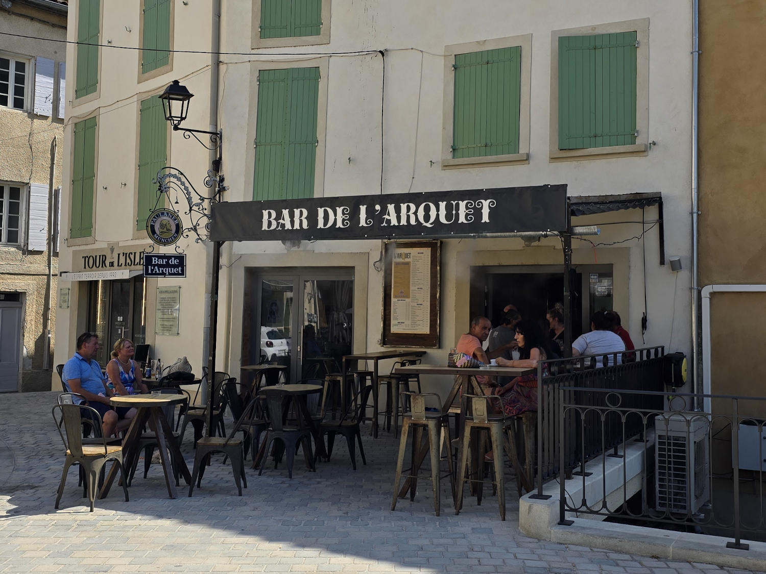 La terrasse ensoleillée d'un café-bar nommé "Bar de l'Arquet", avec plusieurs clients assis à des tables.