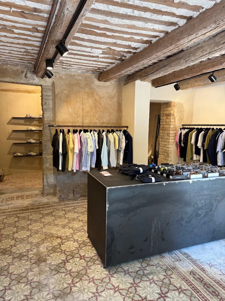 intérieur boutique avec poutres au plafond, carrelage carreaux de ciments usés portants de vetements et ilot central métal noir avec accessoires dessus : casquettes et lunettes