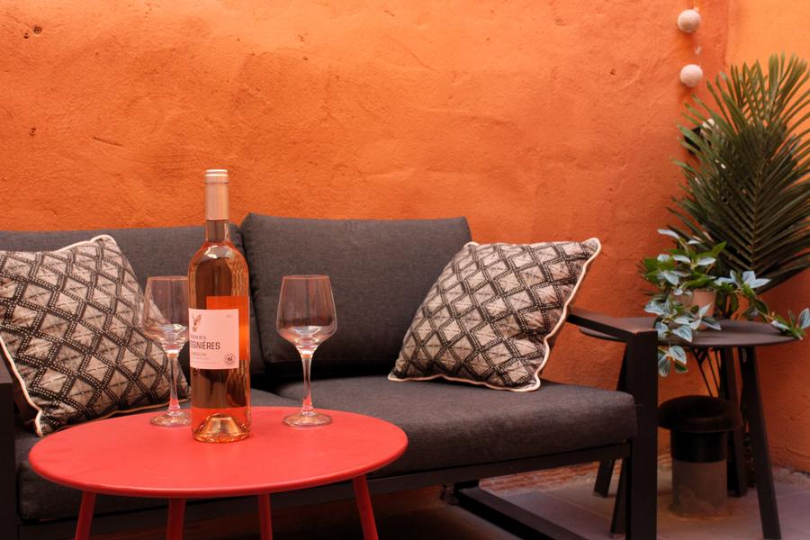 Un petit patio aux murs orangés, chaleureux et intimiste. Un canapé gris foncé avec deux coussins à motifs géométriques noir et blanc occupe le fond de la scène. Devant, une table basse ronde rouge porte une bouteille de vin rosé et deux verres à pied. À droite, une table d’appoint accueille une plante verte en pot, ajoutant une touche de fraîcheur. L’ambiance évoque un moment de détente convivial, parfait pour un apéritif.
